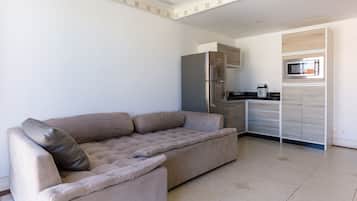 A living room with a beige sofa, a built-in kitchen with a refrigerator and microwave, and a tiled floor.