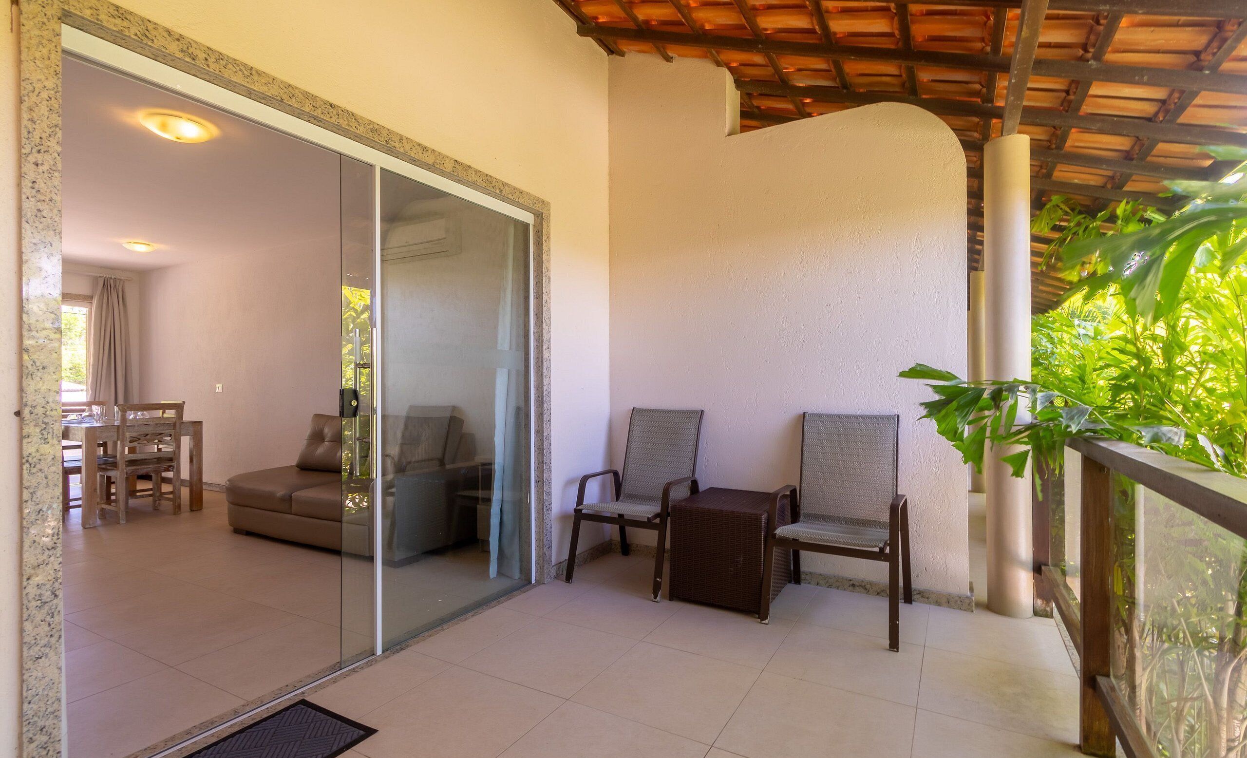 A modern living room with a glass door leading to a dining area, a balcony with wicker chairs, and a view of lush greenery.