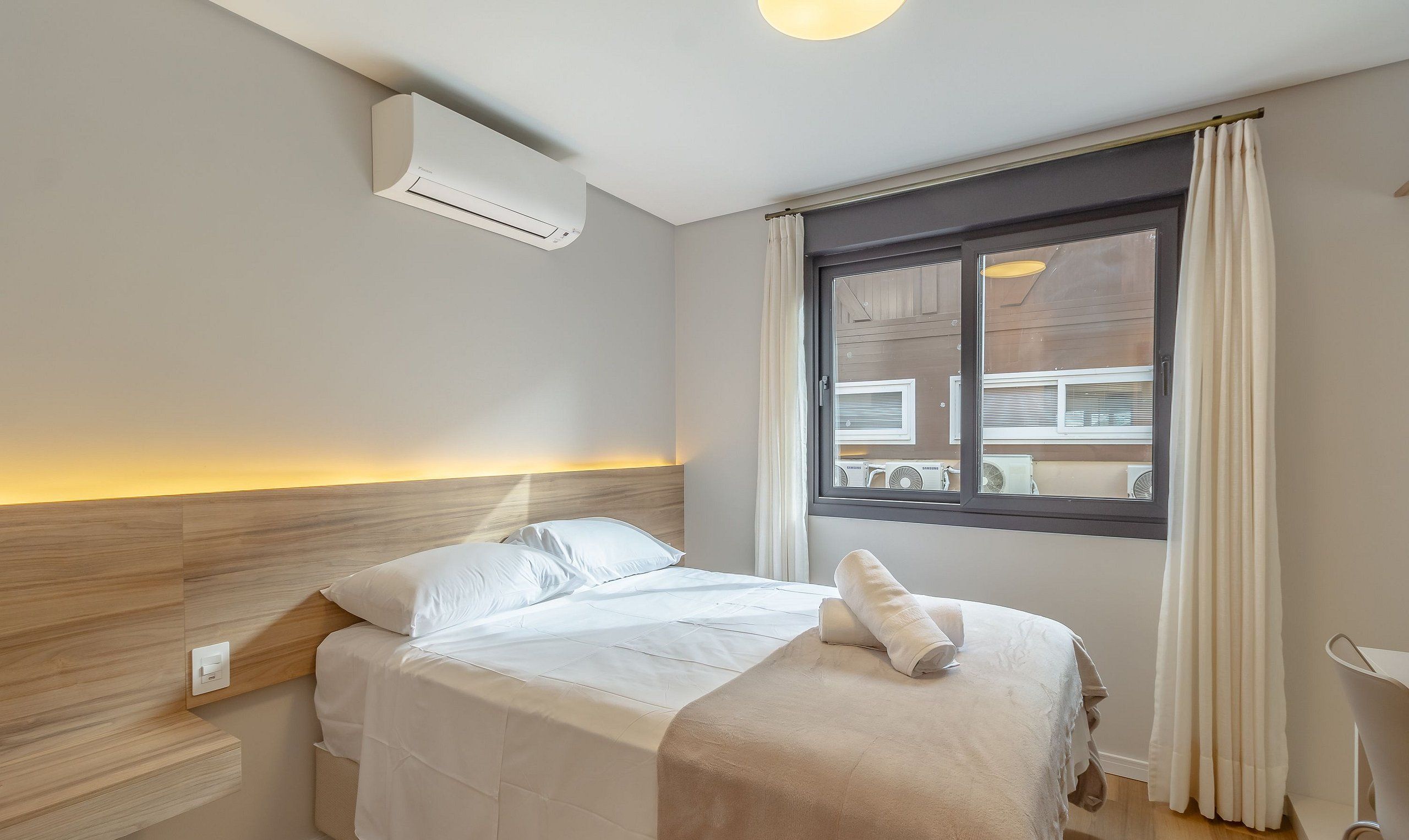 A hotel room with a bed, a window with curtains, a wall-mounted air conditioner, and a bedside table with a lamp.