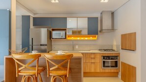 Cocina compacta con microondas, cafetera, estufa a gas y horno empotrado. Hay dos sillas de madera con asientos de mimbre y una mesita con una taza blanca encima.