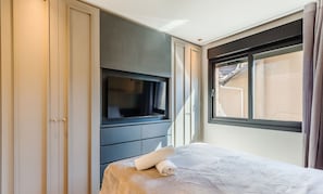 A hotel room with a bed, a TV mounted on a dark-colored cabinet, and a window with curtains. - Apartment in the Center Near Rua Coberta Dlh0303 (Gramado)
