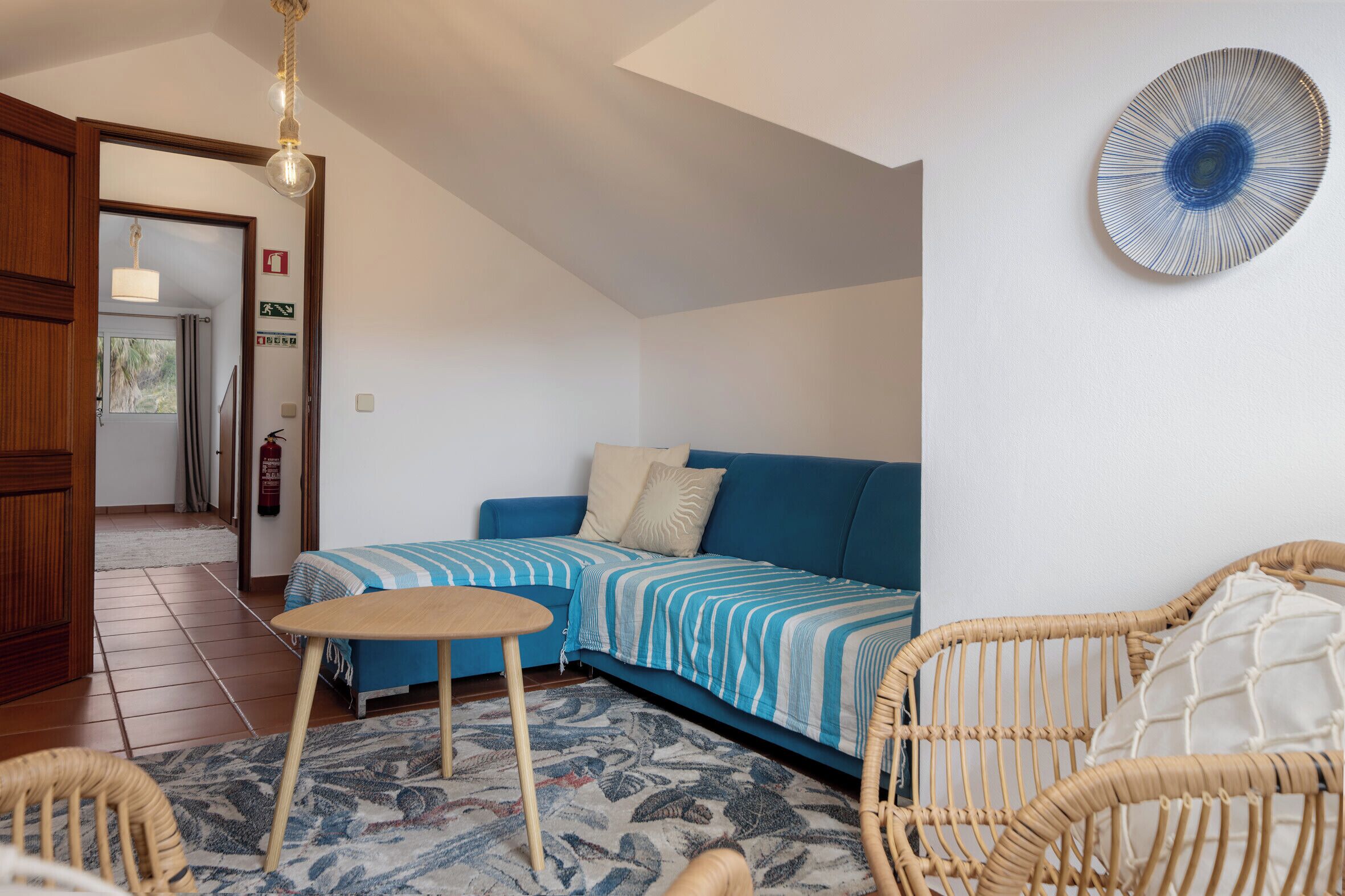 Une pièce avec un canapé bleu, une petite table ronde, des chaises en osier, un tapis à motifs et une assiette décorative accrochée au mur.