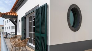 Uma varanda com cadeiras de vime e vista para um edifício com persianas verdes e uma janela circular.