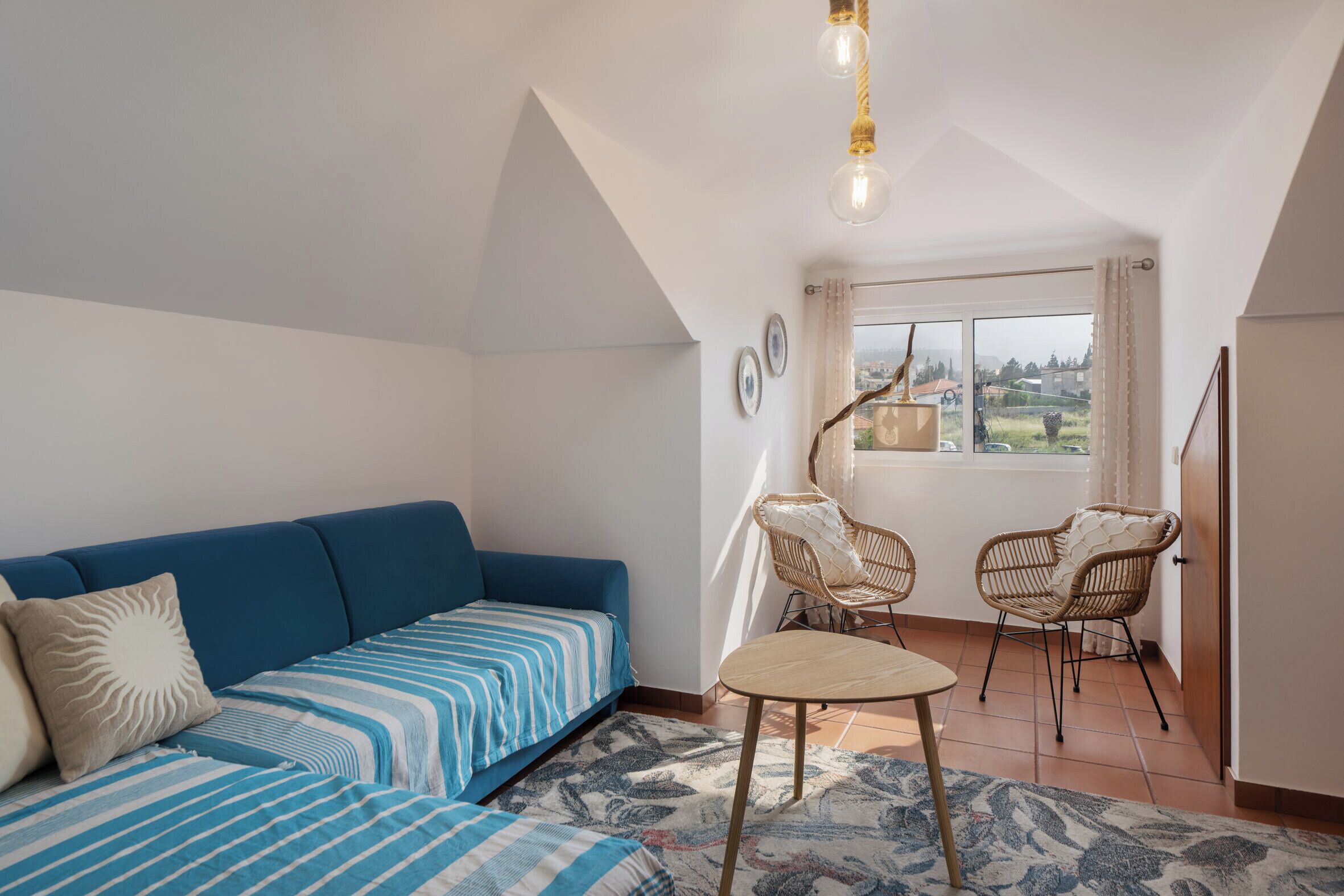 Ein gemütliches Wohnzimmer mit einem blauen Sofa, zwei Korbstühlen, einem kleinen runden Tisch und einem großen Fenster mit Vorhängen.