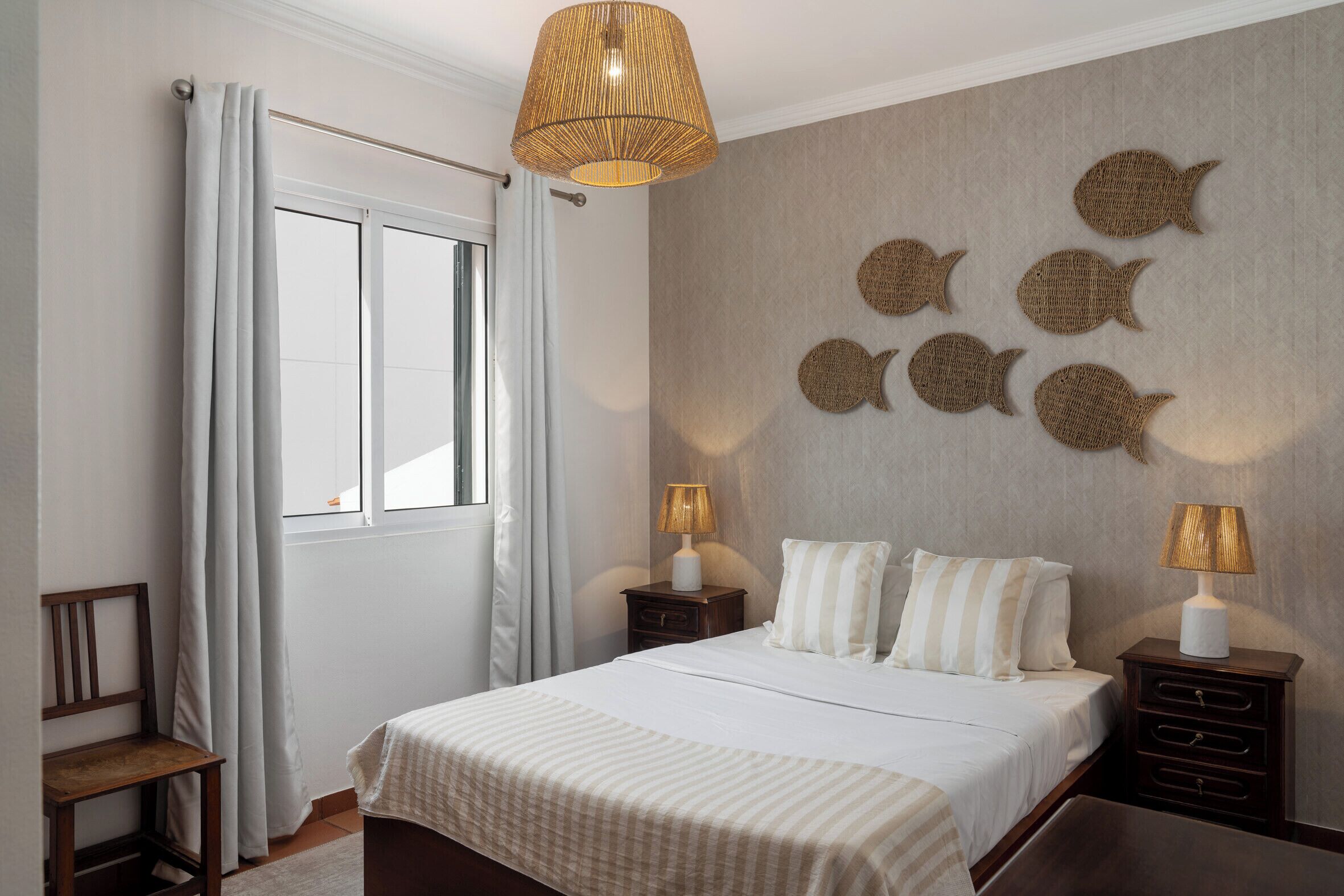 Een slaapkamer met een bed, twee nachtkastjes, een hanglamp, een raam met gordijnen en wanddecoraties.