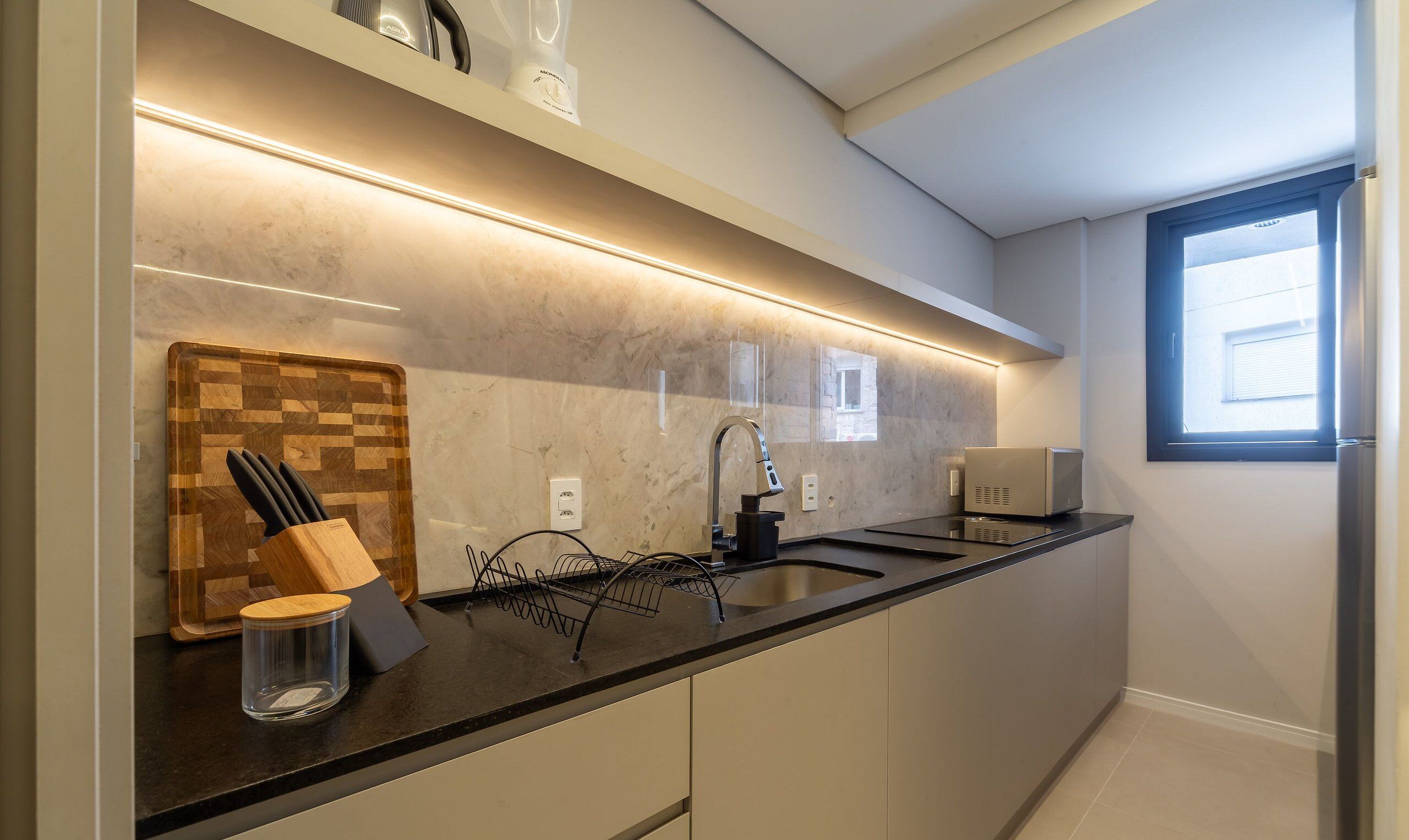 A modern kitchen with a black countertop, a wooden cutting board, a knife block, a glass container, a black faucet, and a microwave.