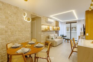 A modern living room with a dining area, featuring a stone wall, wooden furniture, and a built-in kitchen with a white countertop. - Luxurious Apartment in Gramado City Center Dlh0202 (Gramado)