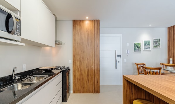 A modern kitchen with a microwave, stove, and wooden panel wall. There is a dining area with a wooden table and chairs. - Charming Apartment in Gramado Dlh0205 (Gramado)