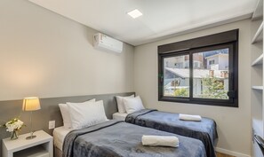 A hotel room with two beds, a window with a view of buildings, a bedside table with a lamp, and an air conditioning unit. - Charming Apartment in Gramado Dlh0205 (Gramado)