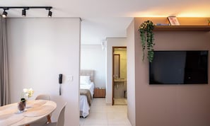 Una habitación de hotel con una cama, un televisor montado en la pared, una pequeña mesa con un jarrón de flores y una planta colgando.