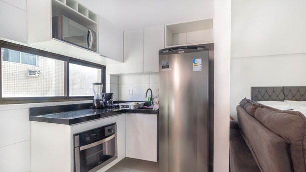 A modern kitchen with a built-in microwave, a stainless steel refrigerator, and a window overlooking a brick wall. - Modern Beachside Comfort in Boa Viagem - Iaa1004 (Recife)