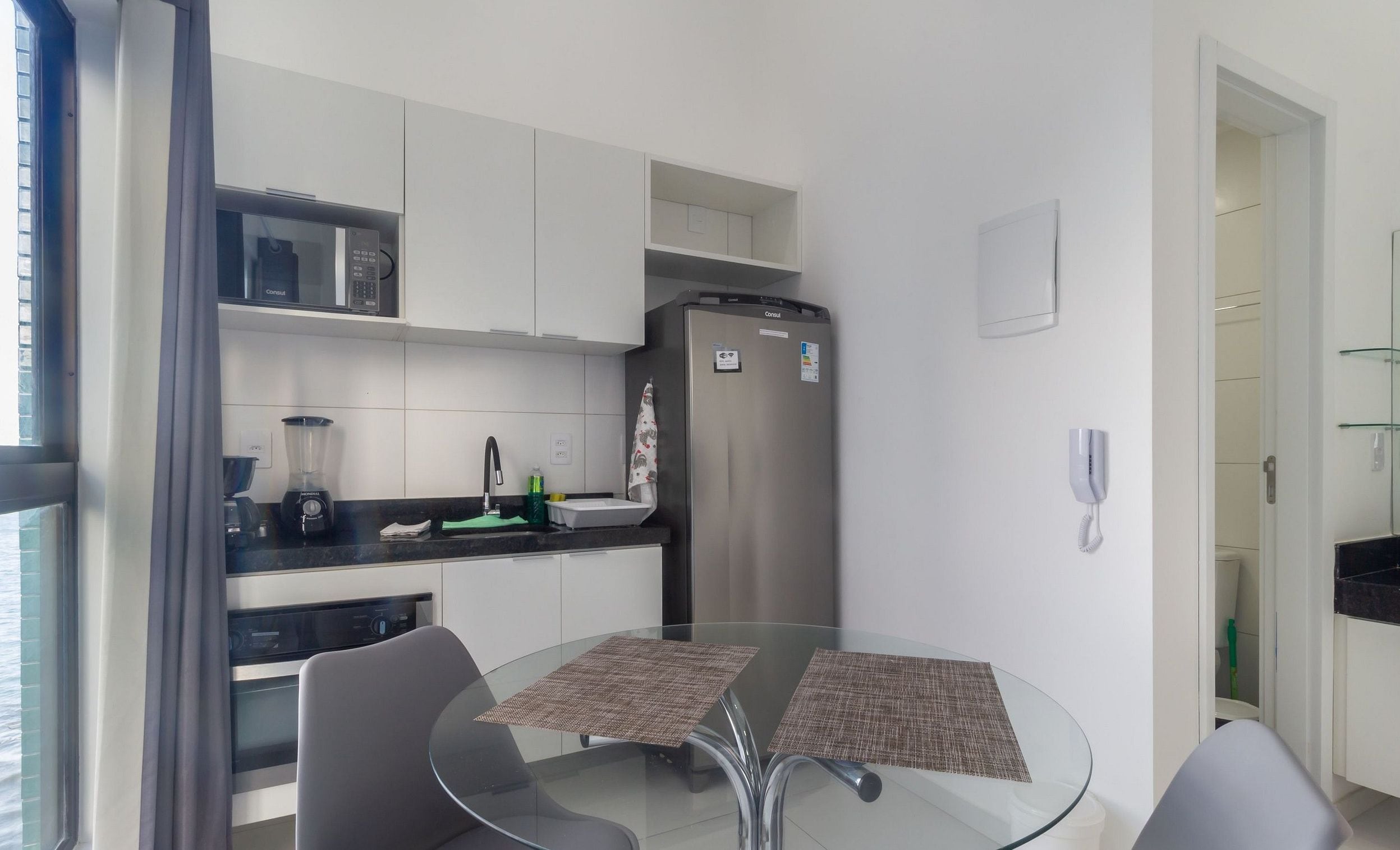 Cozinha moderna com geladeira, micro-ondas e cafeteira. Há uma mesa de vidro com duas cadeiras e uma janela com cortinas.