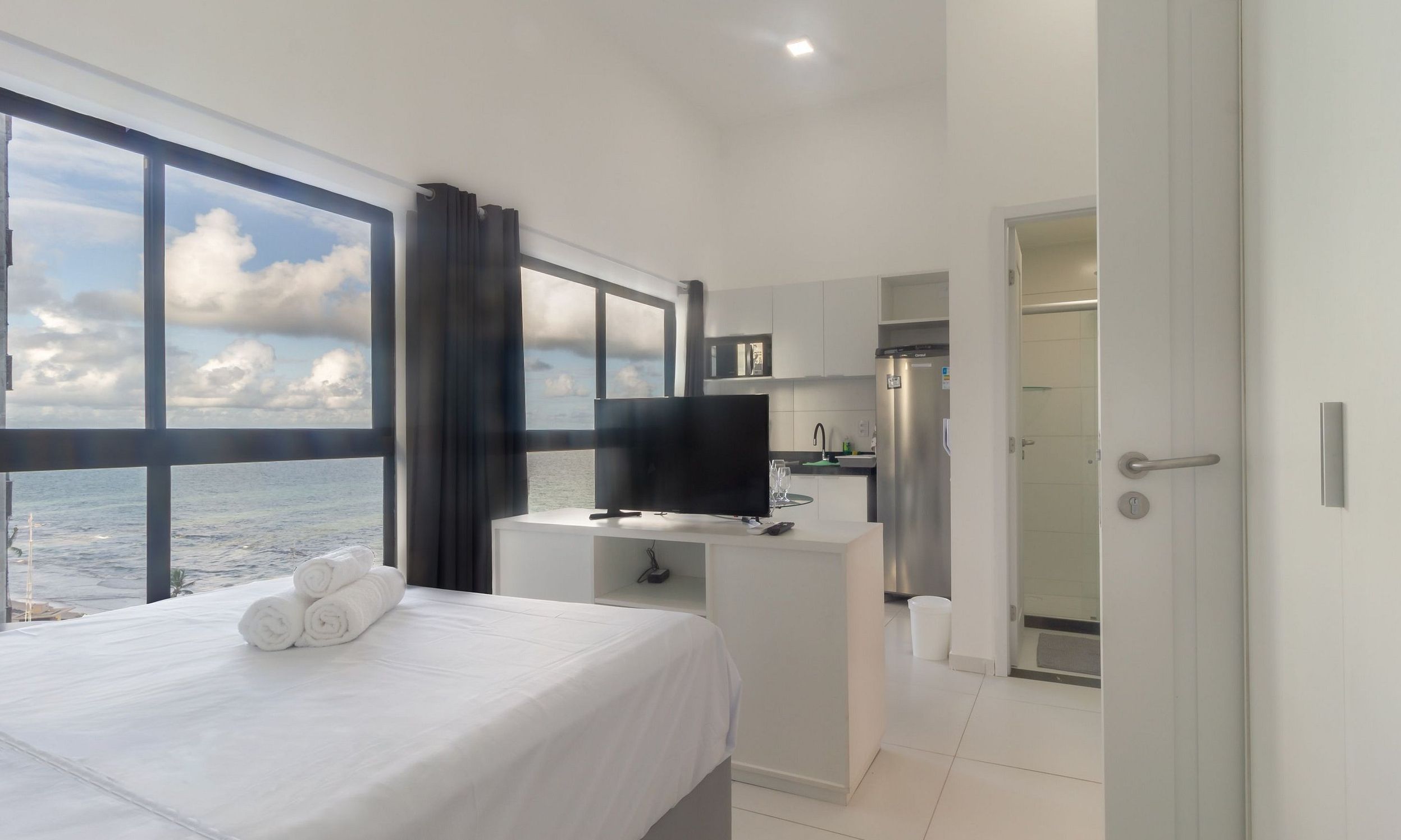 Quarto de hotel com cama, televisão, escrivaninha e vista para o mar.