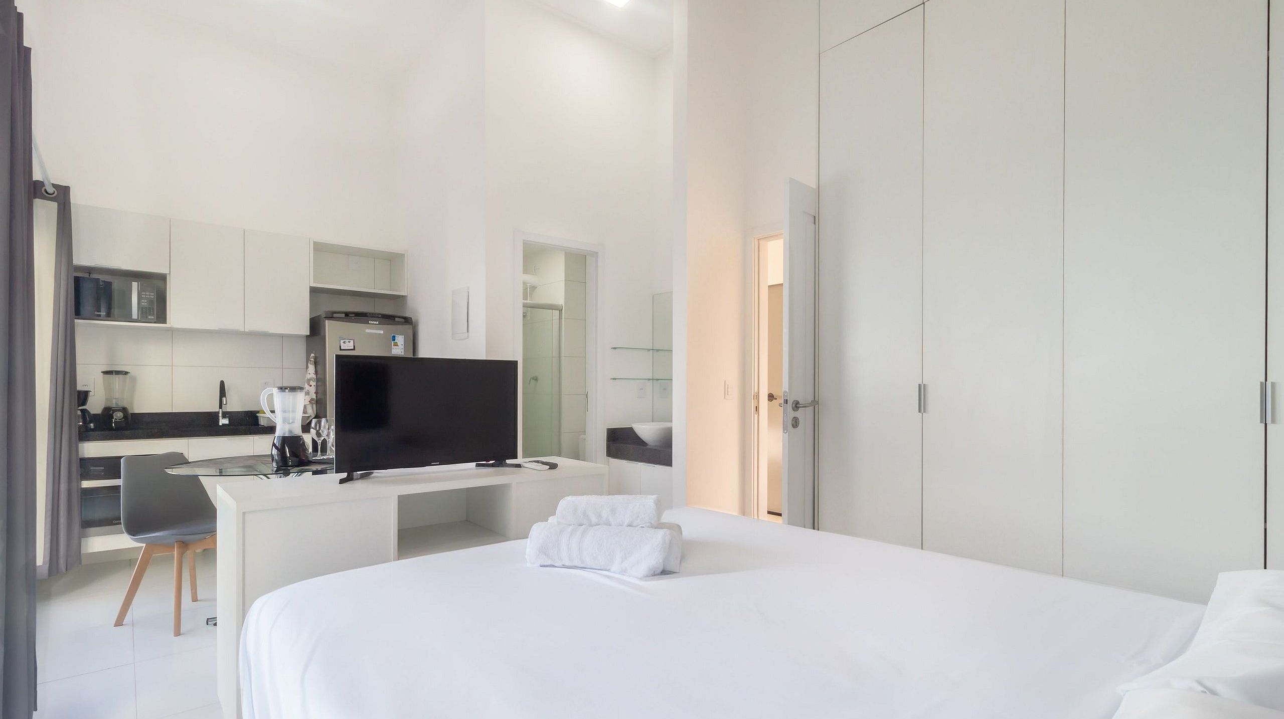 Quarto de hotel com cama, escrivaninha com televisão, cozinha compacta e banheiro.