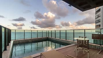 Uma piscina infinita no terraço, com guarda-corpo de vidro, mesa e cadeiras, e vista para o mar e o céu.