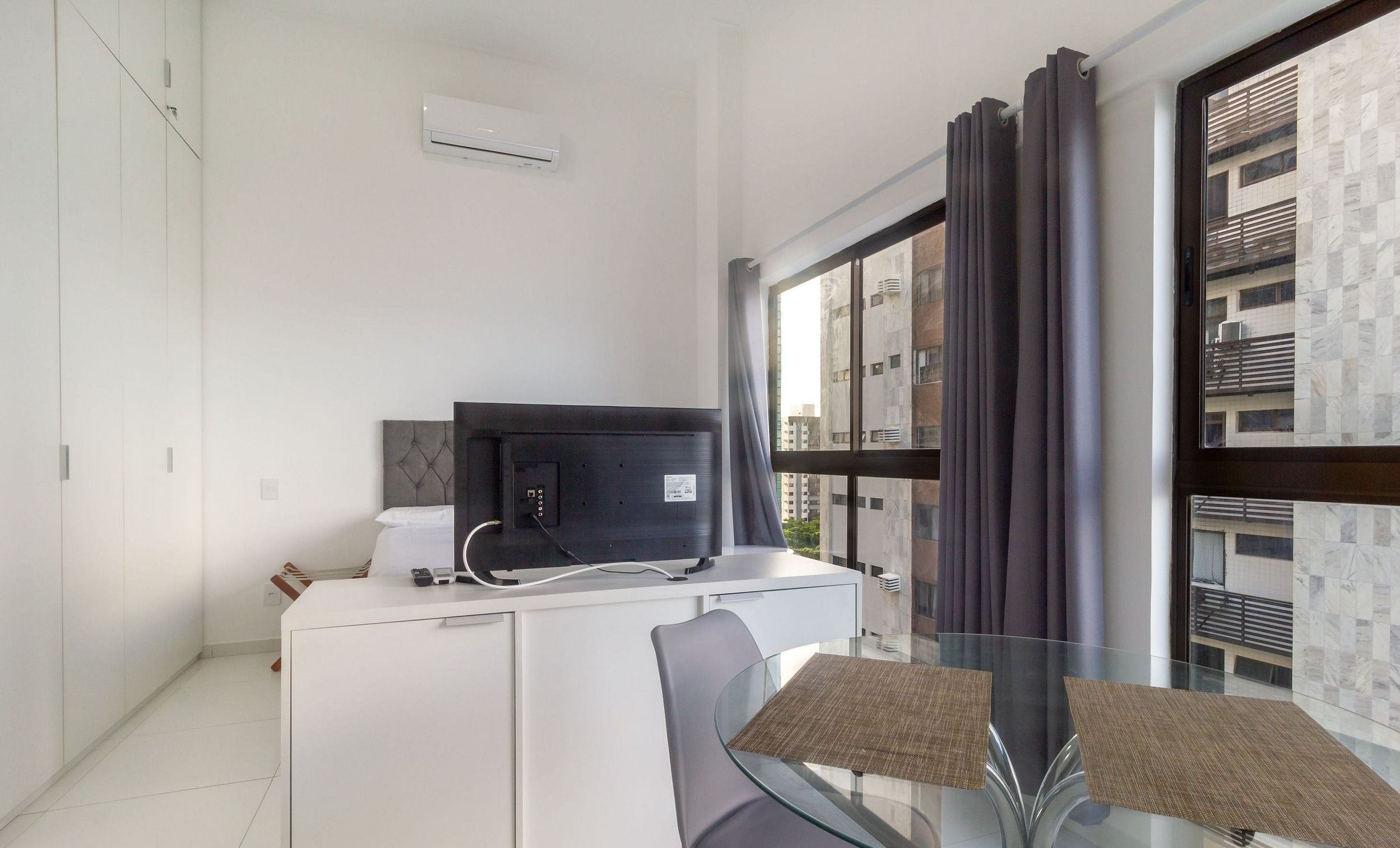 Quarto de hotel com cama, escrivaninha com computador, televisão e janela com cortinas.