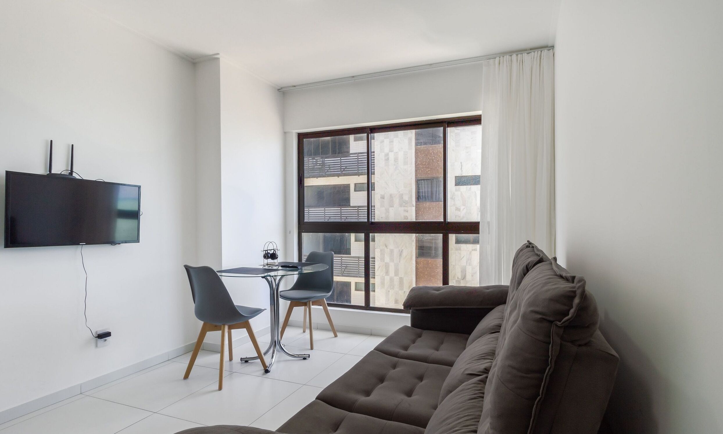 Una sala de estar moderna con un televisor de pantalla plana, una mesa de comedor de vidrio con dos sillas, y un ventanal con vistas a edificios.