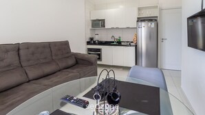 A modern living room with a brown sofa, a glass coffee table, and a remote control. A kitchen area with white cabinets, a microwave, and a refrigerator is visible in the background. - Studio w/ sea View in Bldg w/ gym - Iaa0503 (Recife)