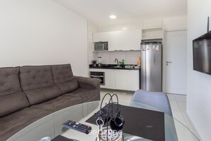 Ein modernes Wohnzimmer mit einem braunen Sofa, einem Glastisch und einer Fernbedienung. Im Hintergrund ist eine Küchenzeile mit weißen Schränken, einer Mikrowelle und einem Kühlschrank zu sehen.