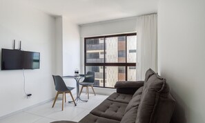 A modern living room with a flat-screen TV, a sofa, a dining table with chairs, and a large window with a view of buildings. - Studio w/ sea View in Bldg w/ gym - Iaa0503 (Recife)