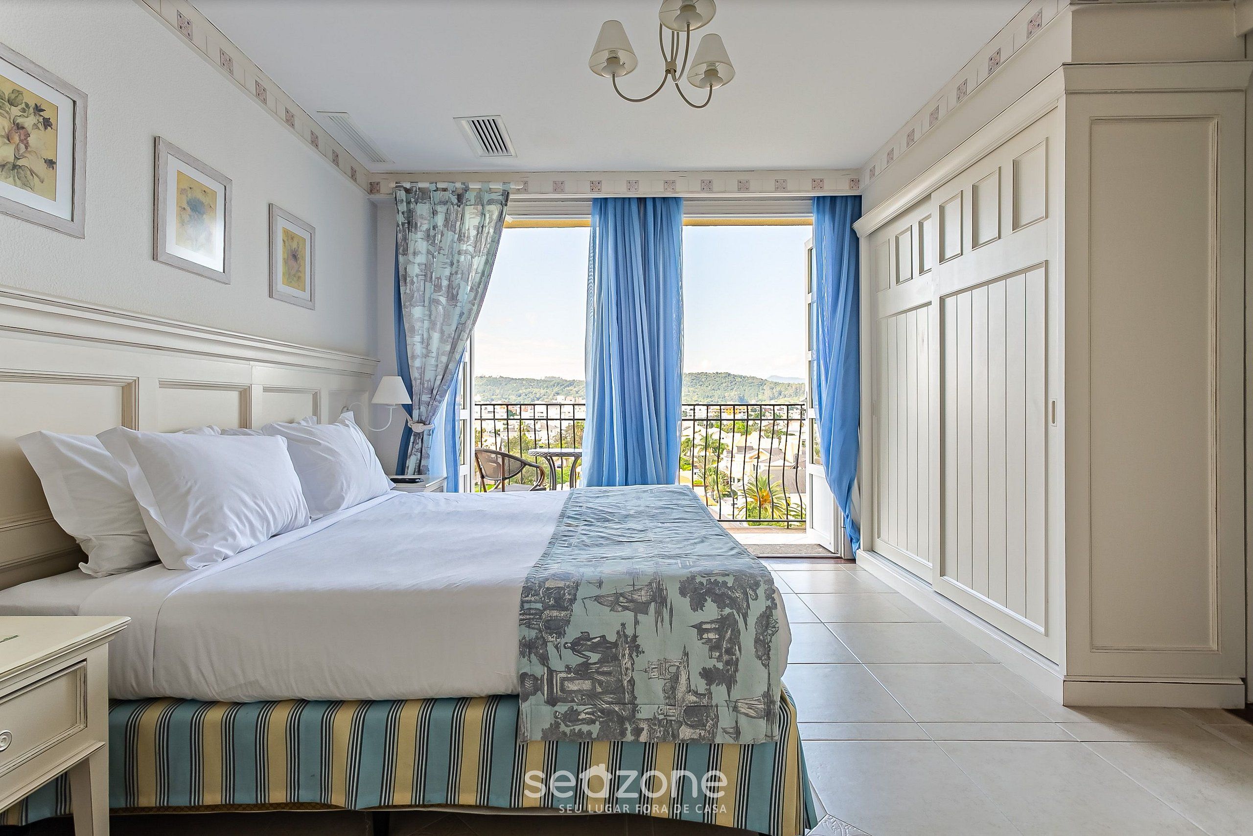 Quarto com cama, mesa de cabeceira, janela com cortinas e vista para o exterior.