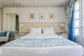 Quarto de hotel com uma cama grande, duas mesas de cabeceira, um criado-mudo, uma cadeira e uma porta com cortina.
