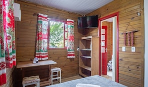 Una habitació d'alta cabana acollidora, amb parets de fusta, un llit, una taula petita amb una cadira, un televisor fixat a la paret i una finestra amb cortines.