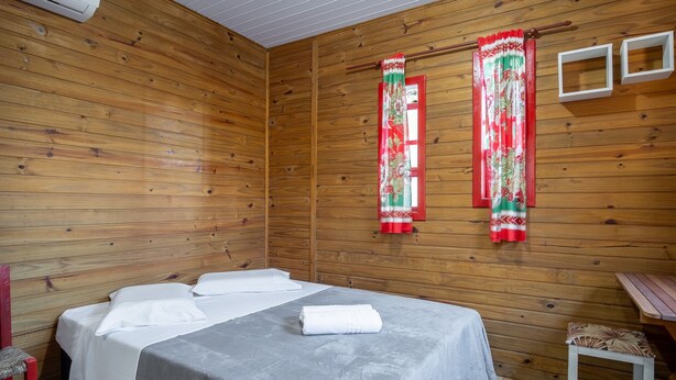 A wooden room with a bed, two windows with red curtains, and a small table. - Charming Cabin 11 Minutes From Rosa Beach Prj001 (Imbituba)