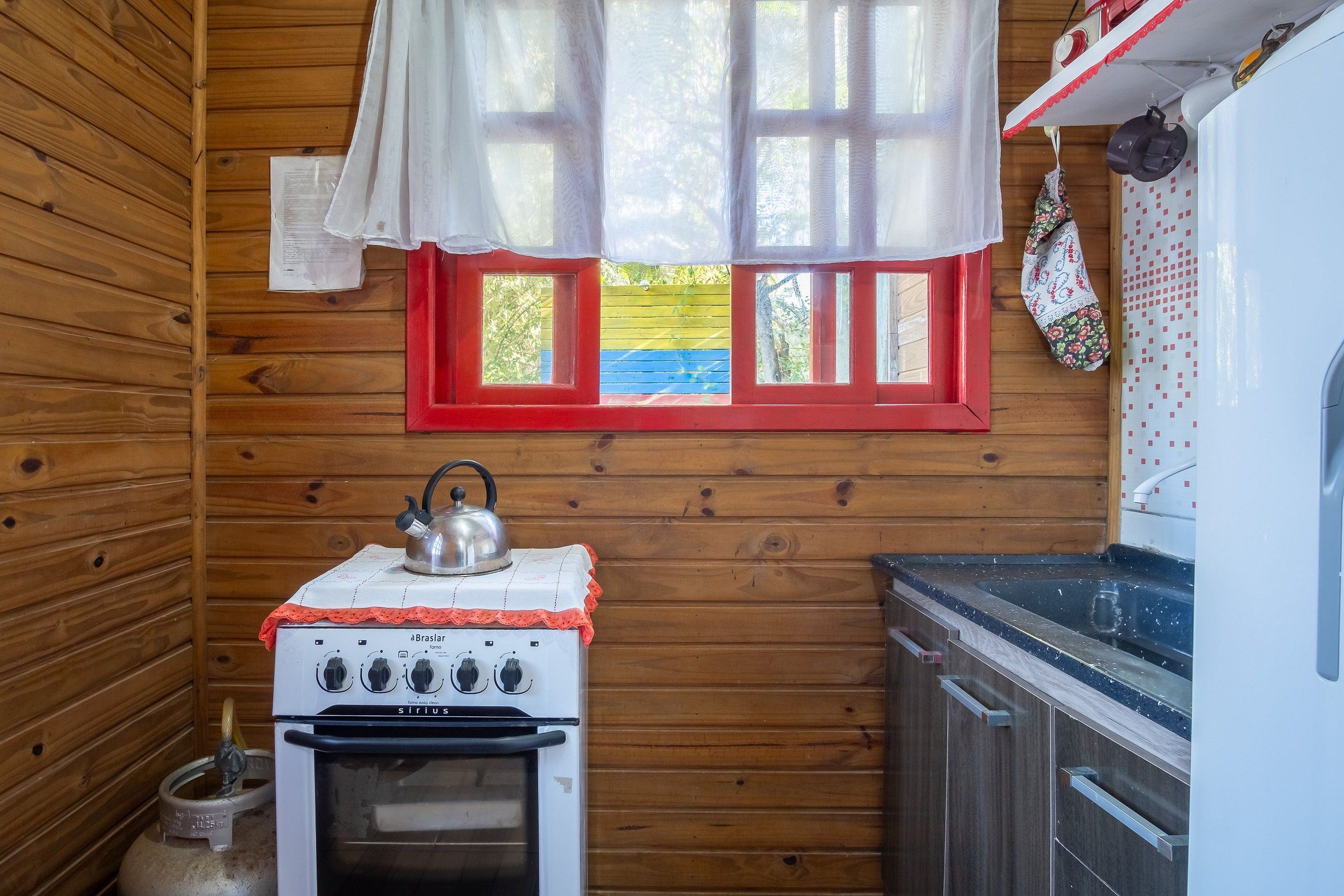 A compact kitchen with a white stove, a kettle, and a window with red frames.
