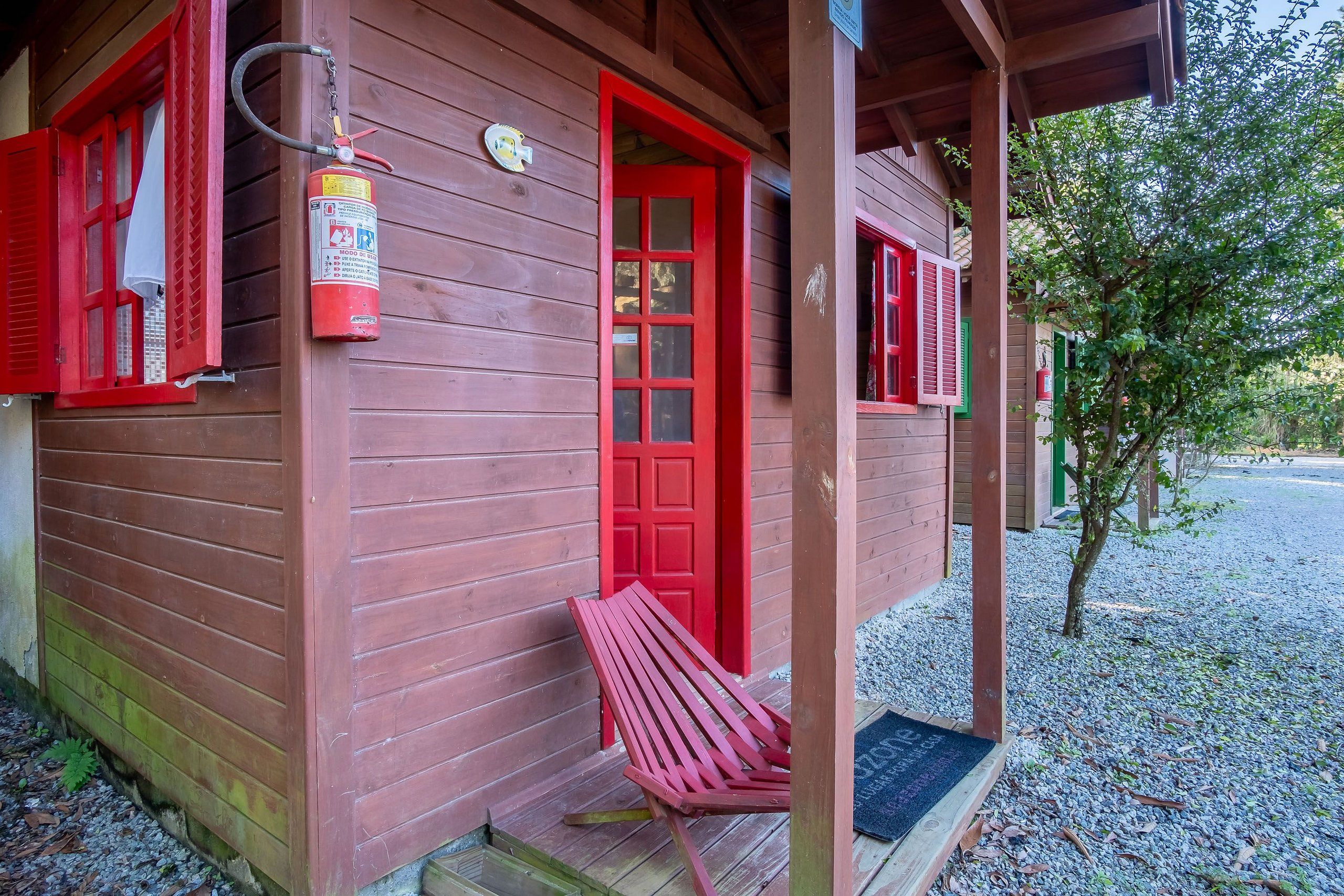 A wooden building with a red door and windows, a fire extinguisher mounted on the wall, and a bench outside.