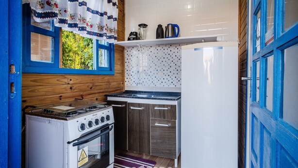 Cocina compacta con estufa, horno y refrigerador. Hay una ventana con cortina y un estante sobre el lavamanos.