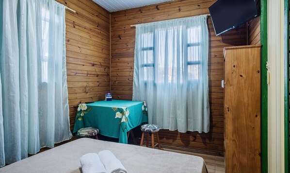A wooden room with a bed, a table with a green tablecloth, a stool, and a television on the wall. - Cozy Cabin in Praia do Rosa - Prj003 (Imbituba)