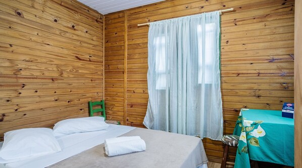 A wooden room with a bed, a green chair, and a table with a blue tablecloth. - Cozy Cabin in Praia do Rosa - Prj003 (Imbituba)