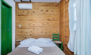 A single bed with white linens, a green door, a wooden wall with a built-in air conditioner, and a green chair. - Cozy Cabin in Praia do Rosa - Prj003 (Imbituba)