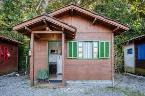 A wooden cabin with a porch, green shutters, and a small kitchen visible through the door. - Cozy Cabin in Praia do Rosa - Prj003 (Imbituba)