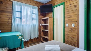 Una habitación con paredes de madera, una mesa con mantel floreado, una cama con manta blanca, un televisor montado en la pared y una puerta con marco verde.