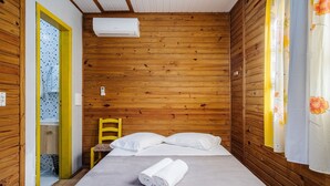 A small, cozy room with wooden walls, a bed with white linens, a chair, and a window with a yellow frame. - Cabin Between Lagoon and Praia do Rosa - Prj004 (Imbituba)