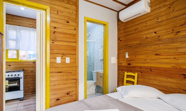 Quarto compacto com paredes de madeira, uma cama, um banheiro pequeno com vaso sanitário e chuveiro, e uma cozinha compacta com fogão.