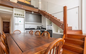 Een moderne keuken met een houten trap, een eethoek met een grote tafel en stoelen, en uitzicht op buiten.
