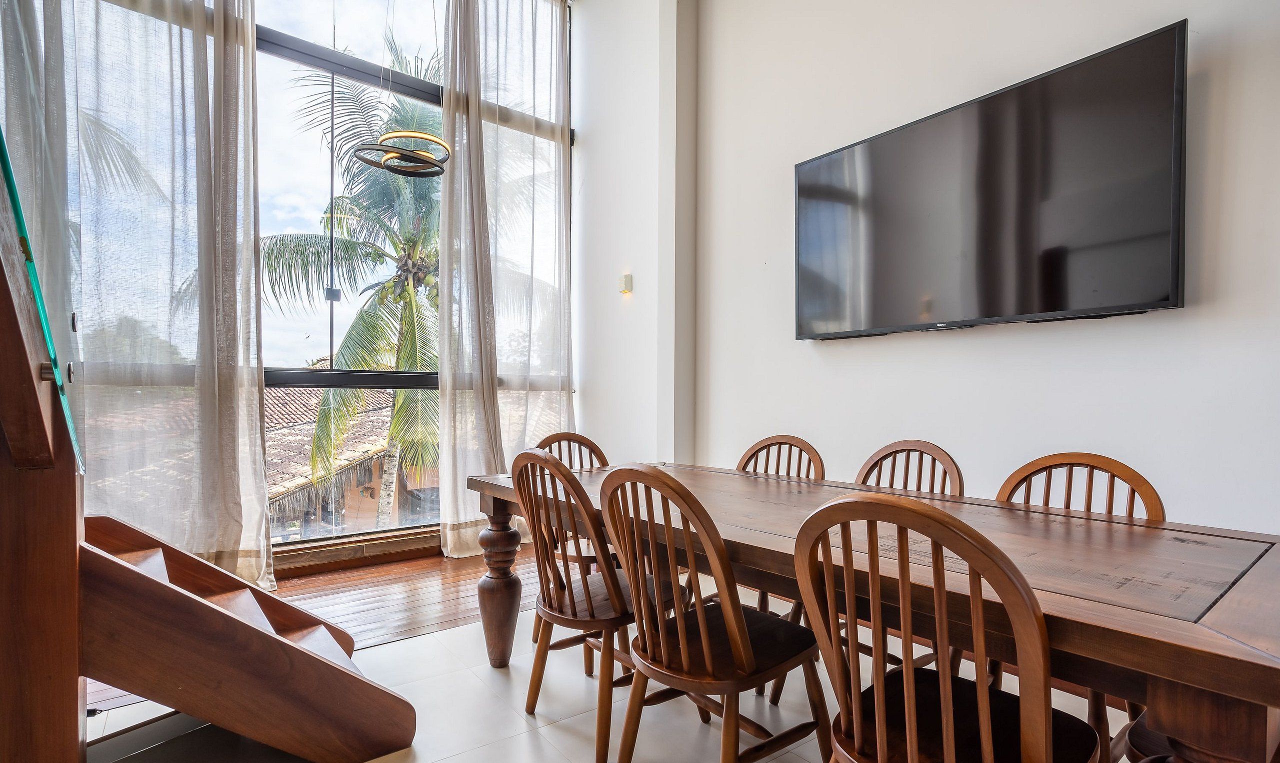 Uma área de refeições com mesa e cadeiras de madeira, uma televisão de tela plana grande fixada na parede e vista para palmeiras através de amplas janelas.