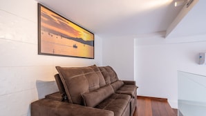 A modern living room with a brown sofa, a large framed sunset painting, and a white tiled wall. - Two-story Apt 200m From Barra Grande Beach Cpr0207 (Maraú)