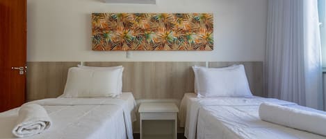 A hotel room with two beds, a small bedside table, a wall-mounted air conditioner, and a tropical leaf-patterned artwork.