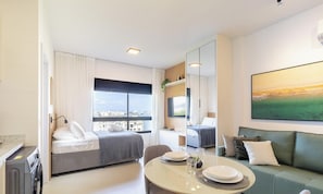 Habitació d'hotel moderna amb llit, taula de menjador amb plats i coberts, sofà, finestra gran amb vista a la ciutat i un quadre a la paret.