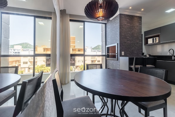 Uma zona de refeições moderna com uma grande mesa redonda e cadeiras, uma parede preta com forno embutido e janelas amplas com vista para a cidade.