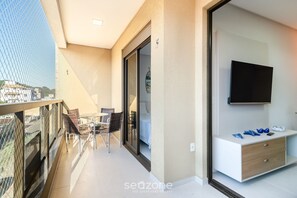 Un balcón con baranda de vidrio, una zona de comedor con sillas, una puerta corrediza de vidrio que da a un dormitorio y un televisor montado en la pared.