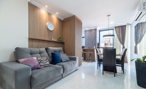 A modern living room with a grey sofa, a dining area with a table and chairs, and a kitchen in the background.