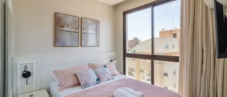 A bedroom with a large window, a bed with pink bedding, and artwork on the wall.