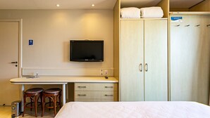 Quarto de hotel com uma cama, uma escrivaninha com dois banquinhos, uma televisão e um guarda-roupa com espelho.