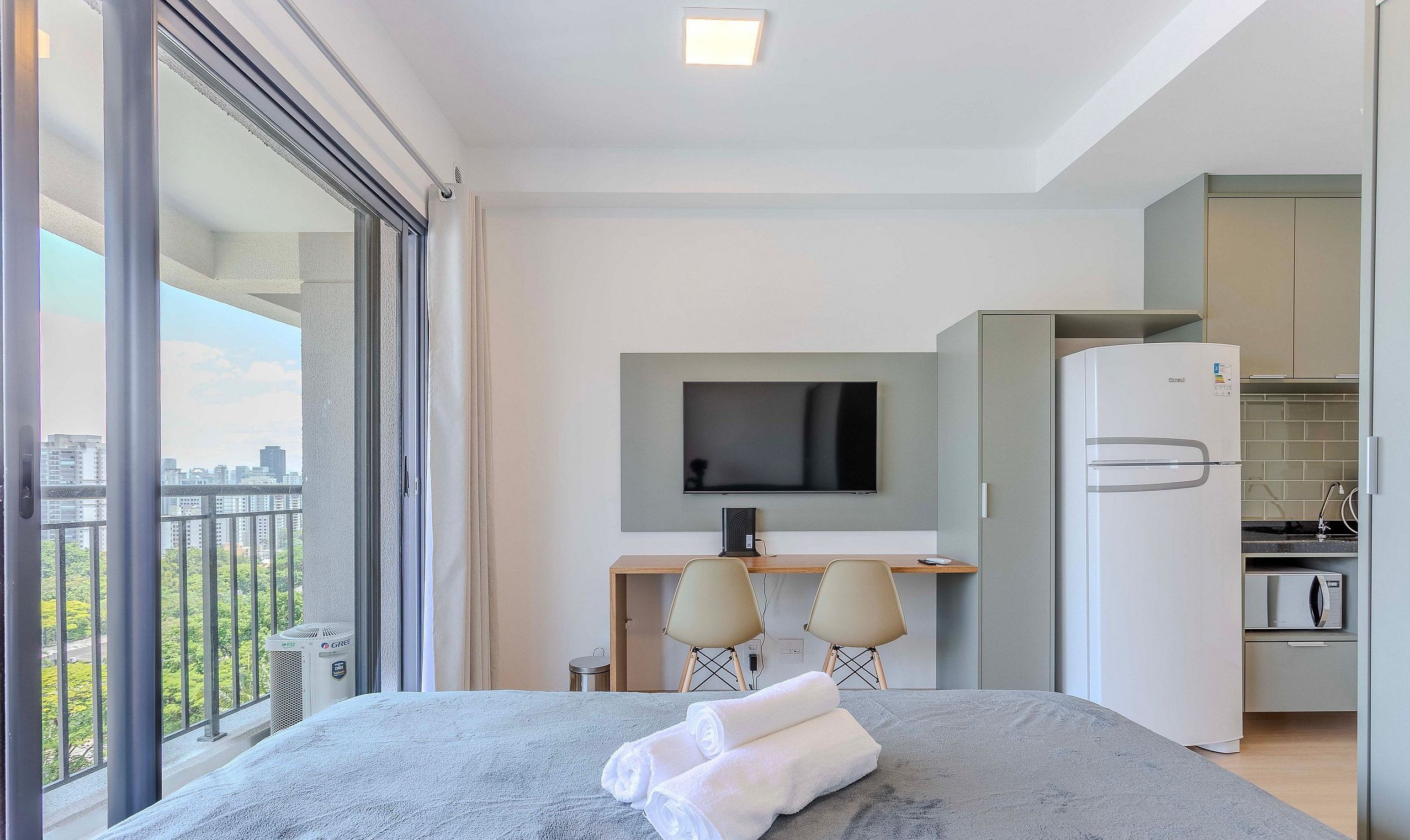 Ein modernes Hotelzimmer mit Balkon, einem Bett mit Handtüchern, einem Schreibtisch mit Stühlen, einem Fernseher und einer Küchenzeile.