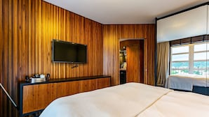 Quarto de hotel com parede de painel de madeira, televisão de tela plana fixada acima de um armário de madeira, cama grande com roupa de cama branca e janela com cortinas.